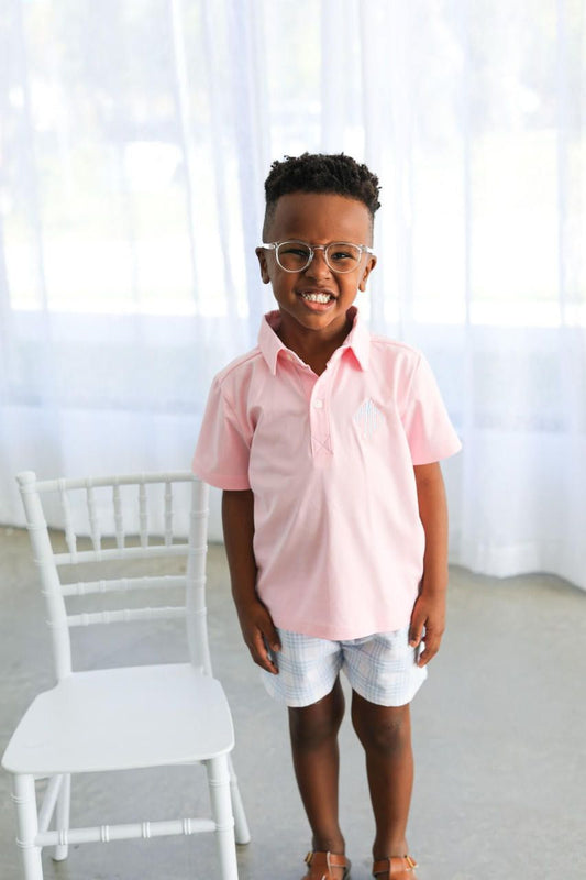 Child wearing a pink shirt and glasses standing in a bright room with white curtains.
