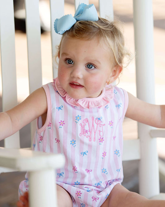 Child wearing a pink sleeveless bubble with floral patterns and a blue bow in hair.