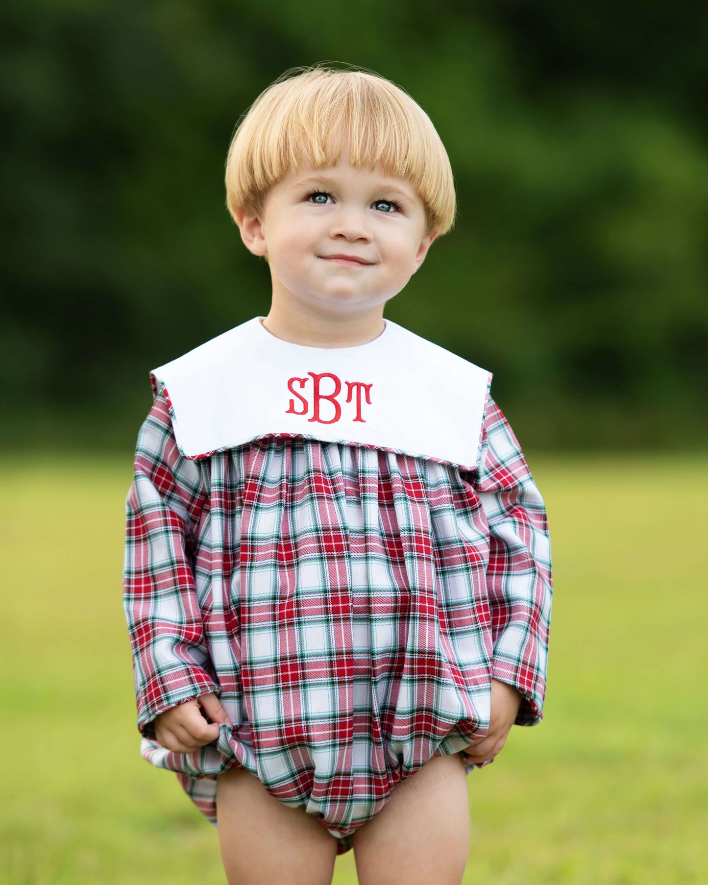 Child wearing a plaid romper with a white collar and red monogram letters outdoors.