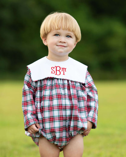 Child wearing a plaid romper with a white collar and red monogram letters outdoors.