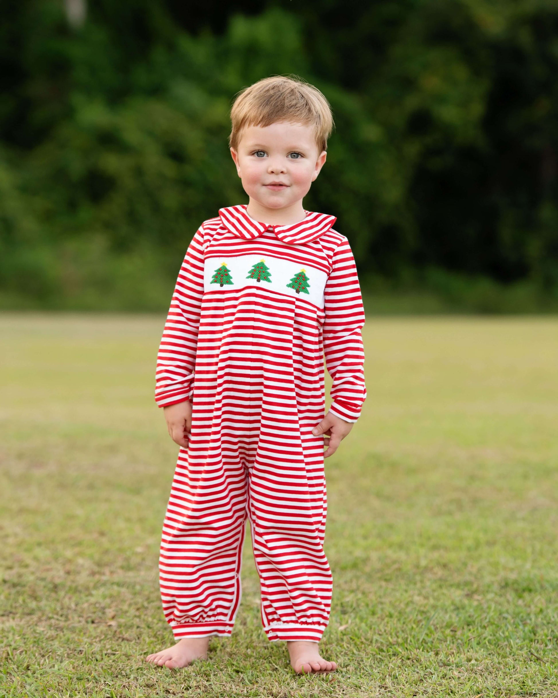 Child wearing a red and white striped onesie with a smocked Christmas tree design outdoors.