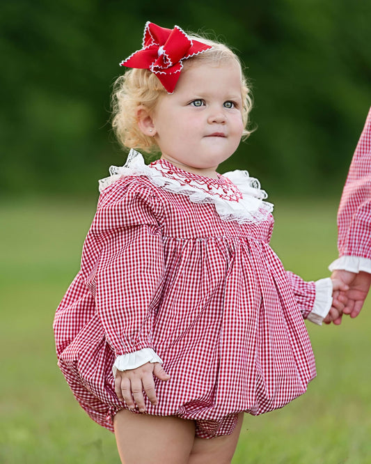 Child wearing a red checkered dress with a white lace trim and a red bow in hair, standing outdoors.