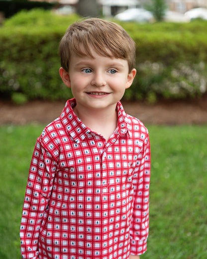 Child wearing a red checkered shirt standing outdoors with greenery in the background