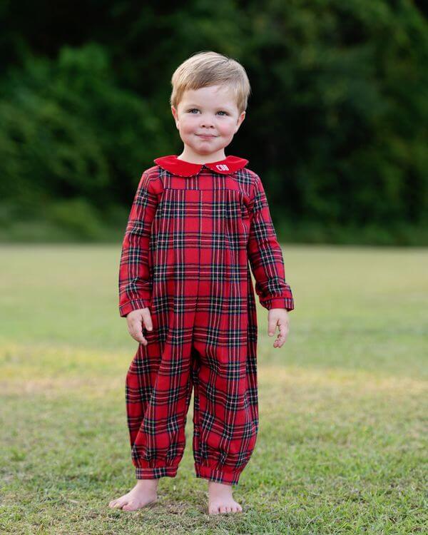 Child wearing a red plaid long romper standing on grass with a blurred green background