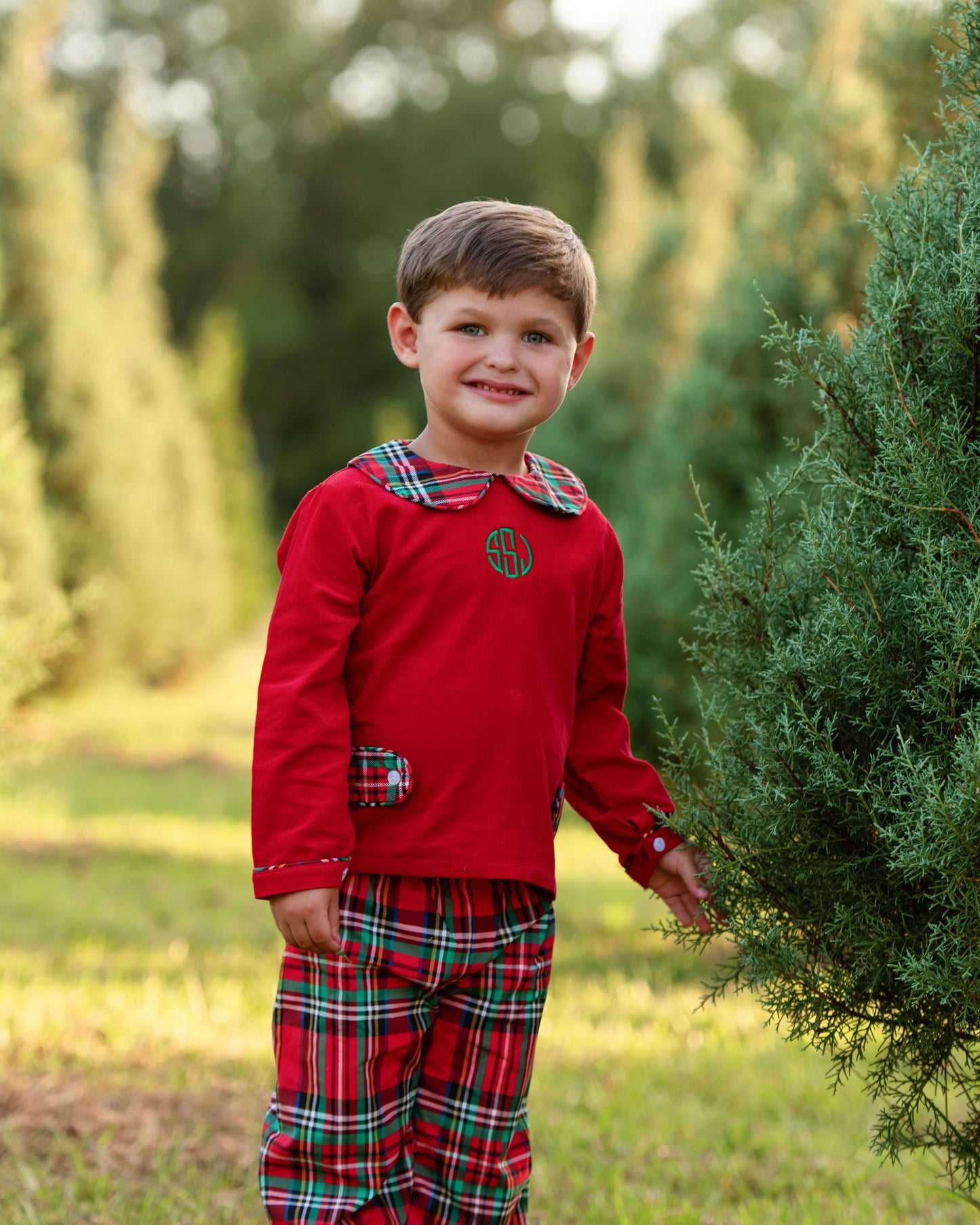 Child wearing a red plaid outfit with a monogram, standing outdoors near greenery.