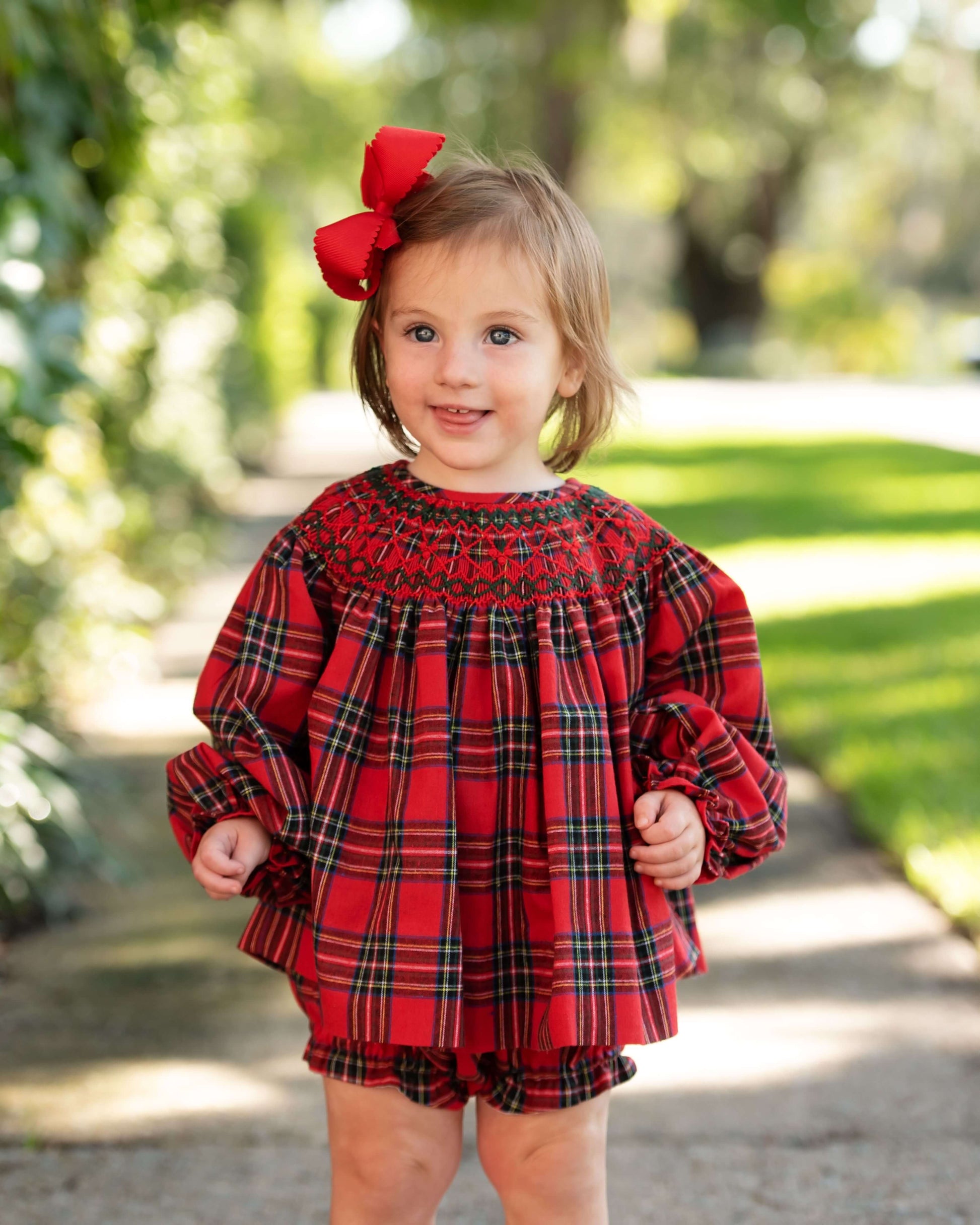 Child wearing a red plaid smocked diaper set with a large bow, standing outdoors