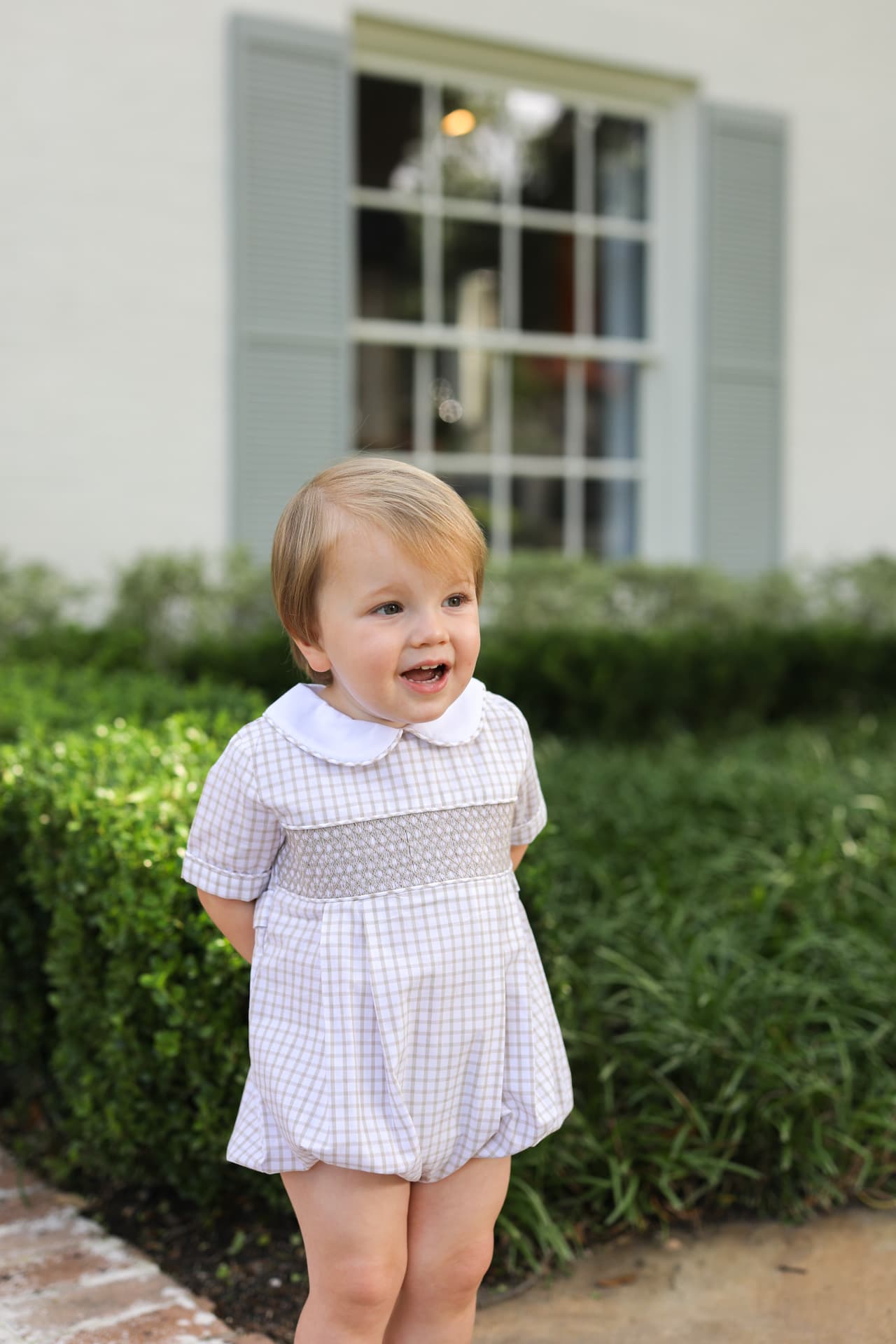 Child wearing a smocked khaki bubble standing in front of a house with greenery.