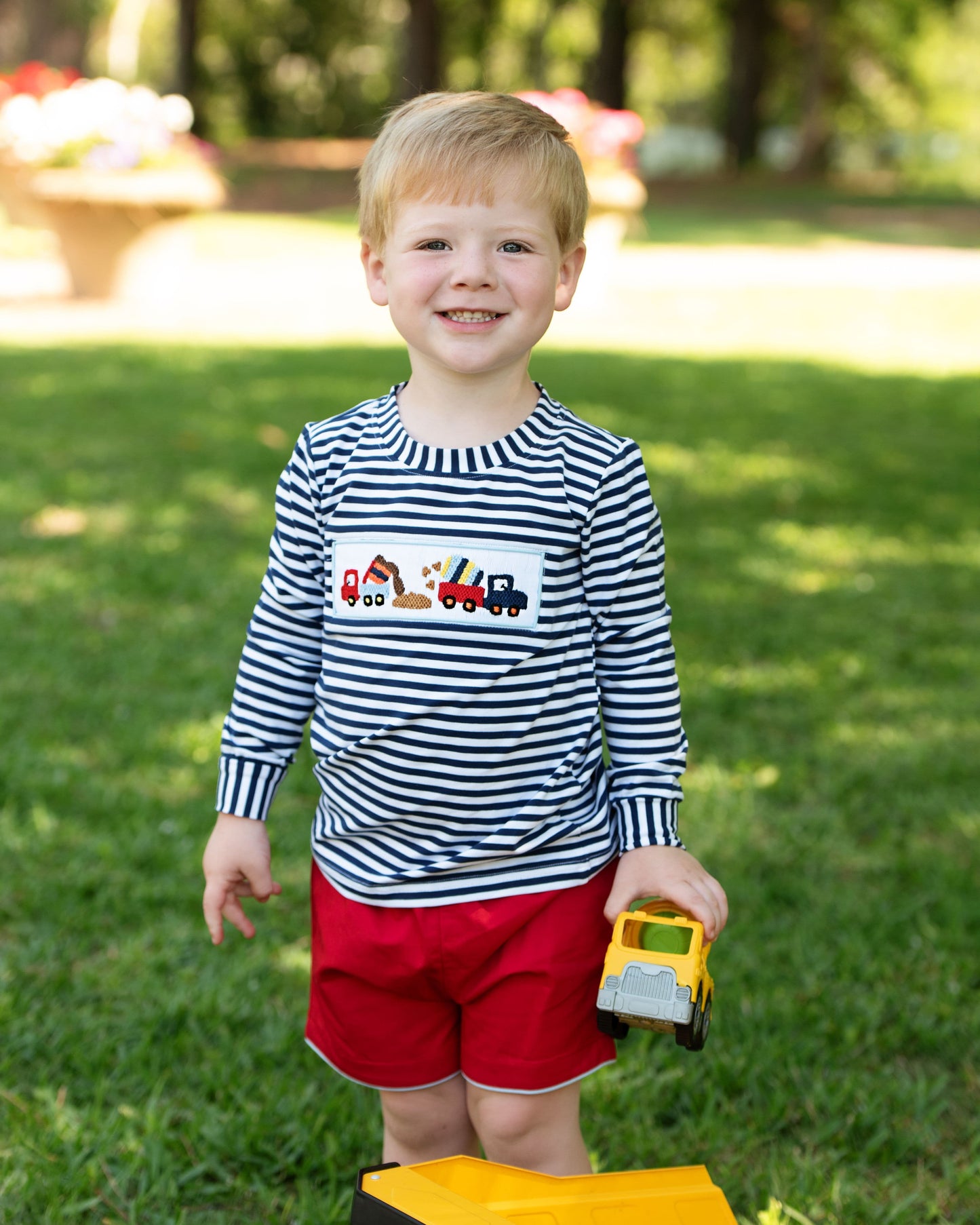 Child wearing a striped shirt with vehicle graphics and red shorts holding a toy truck outdoors