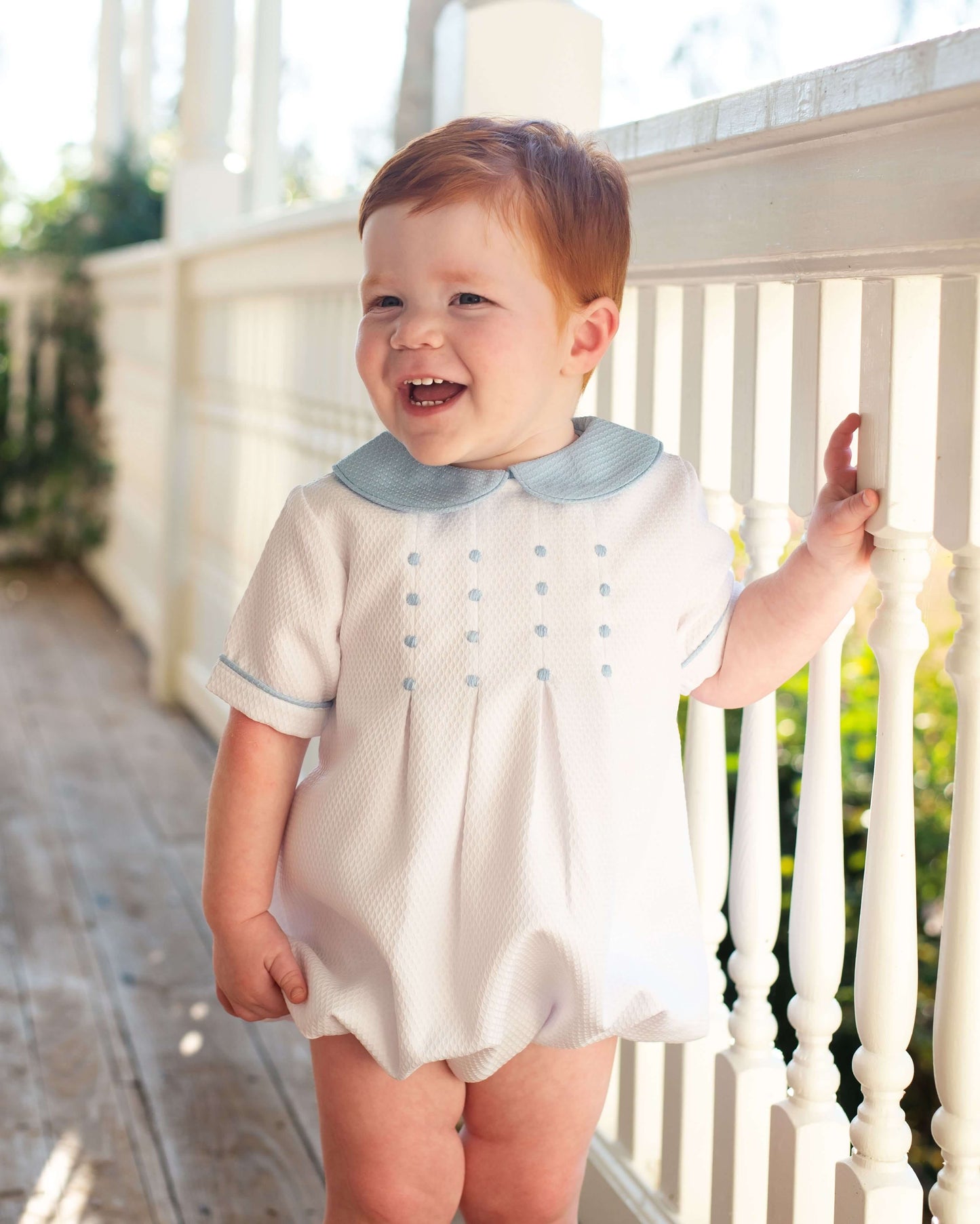 Child wearing a white bubble with blue trim and embroidery on a porch