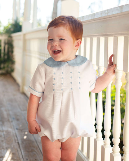 Child wearing a white bubble with blue trim and embroidery on a porch
