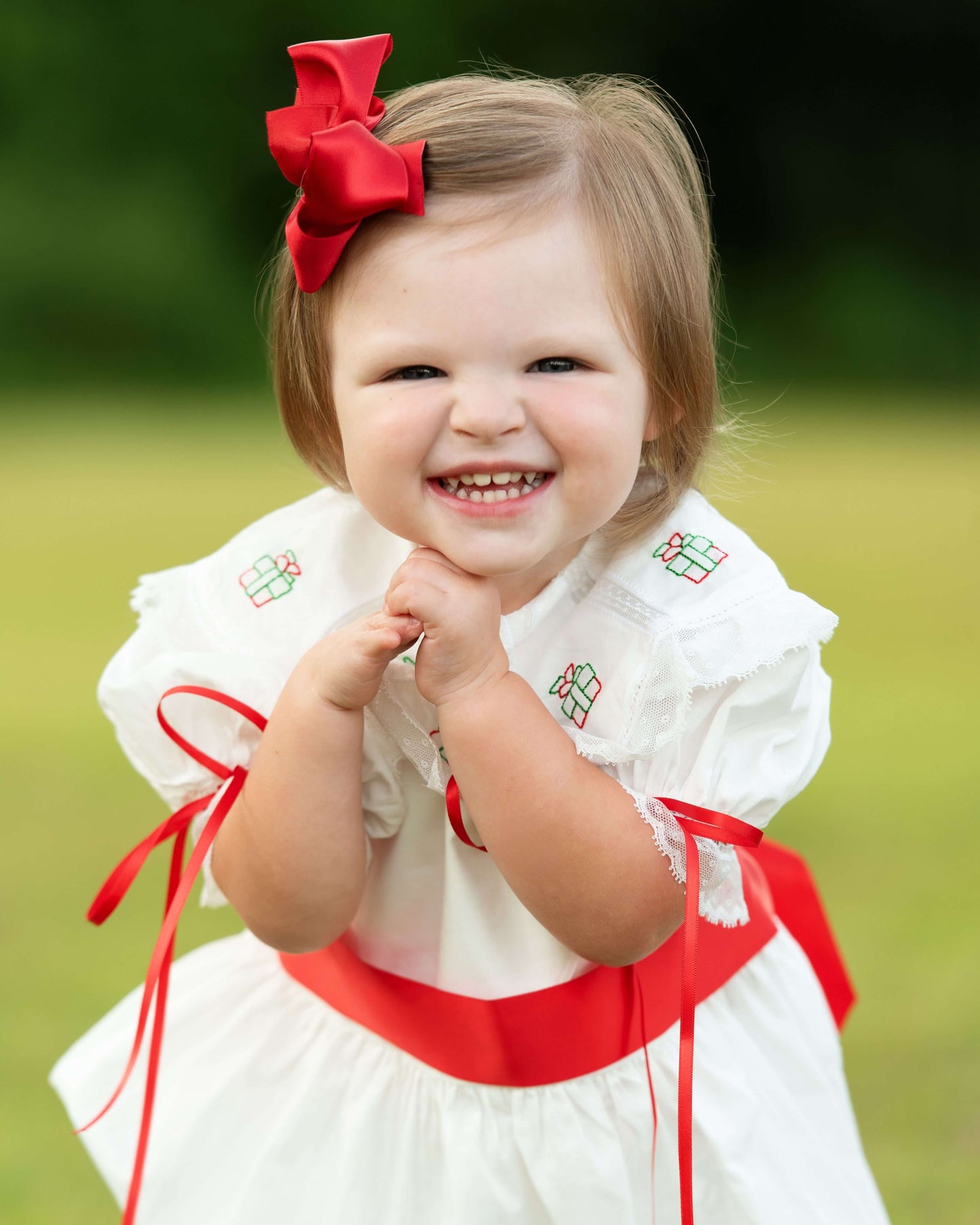 Child wearing a white dress with red accents and a red bow in a grassy outdoor setting
