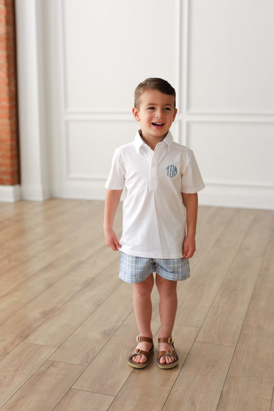 Child wearing a white polo shirt with a monogram and checkered shorts standing on a wooden floor.