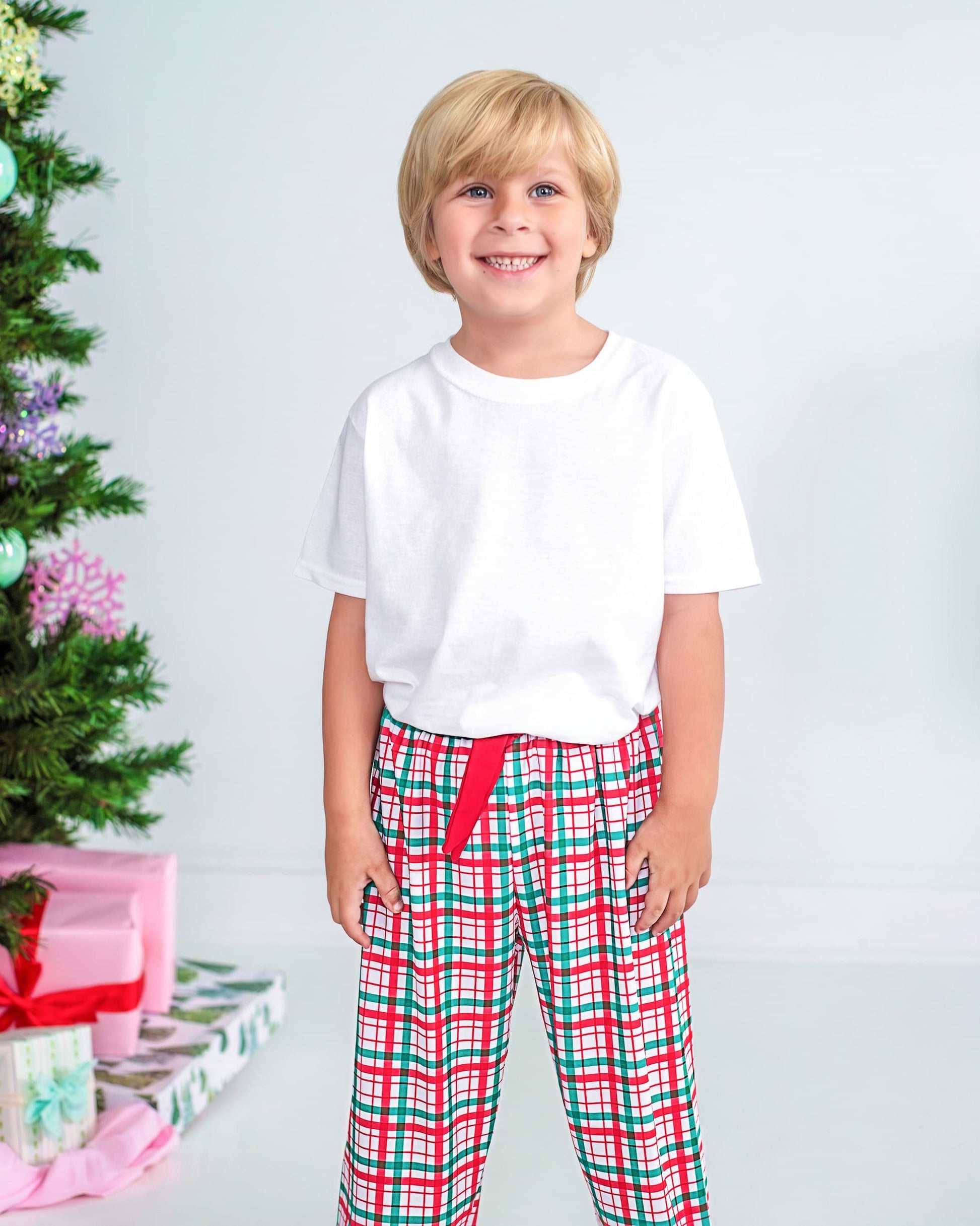 Child wearing a white shirt and red and green plaid pants standing in front of a Christmas tree with presents.