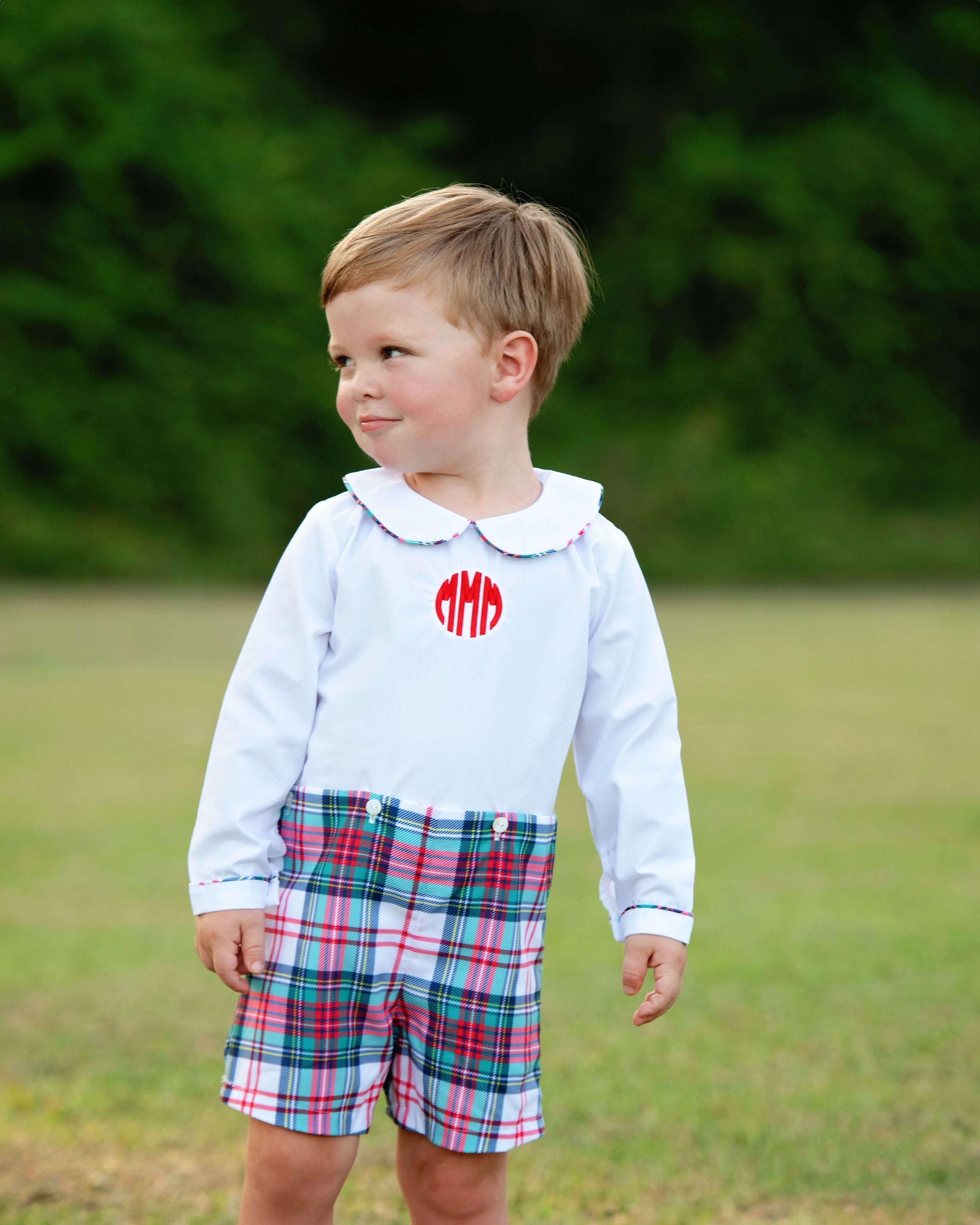 Child wearing a white shirt with red monogram and button-on plaid shorts outdoors