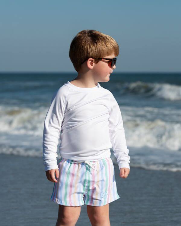 Child wearing sunglasses and a white long-sleeve shirt with colorful shorts standing on a beach.