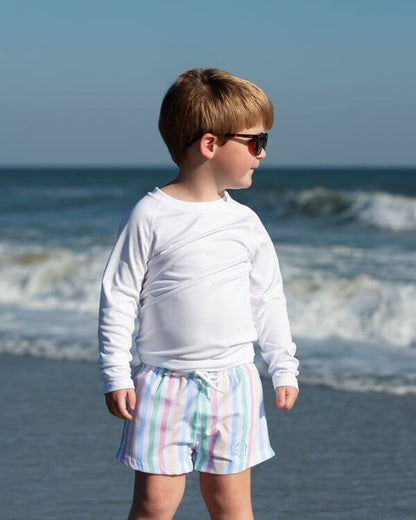 Child wearing sunglasses and a white long-sleeve shirt with colorful shorts standing on a beach.