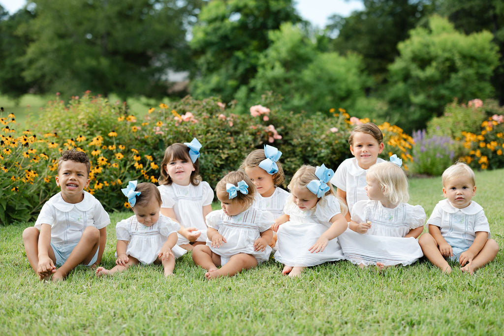 Children in white outfits with blue bows sitting on grass with flowers and trees in the background