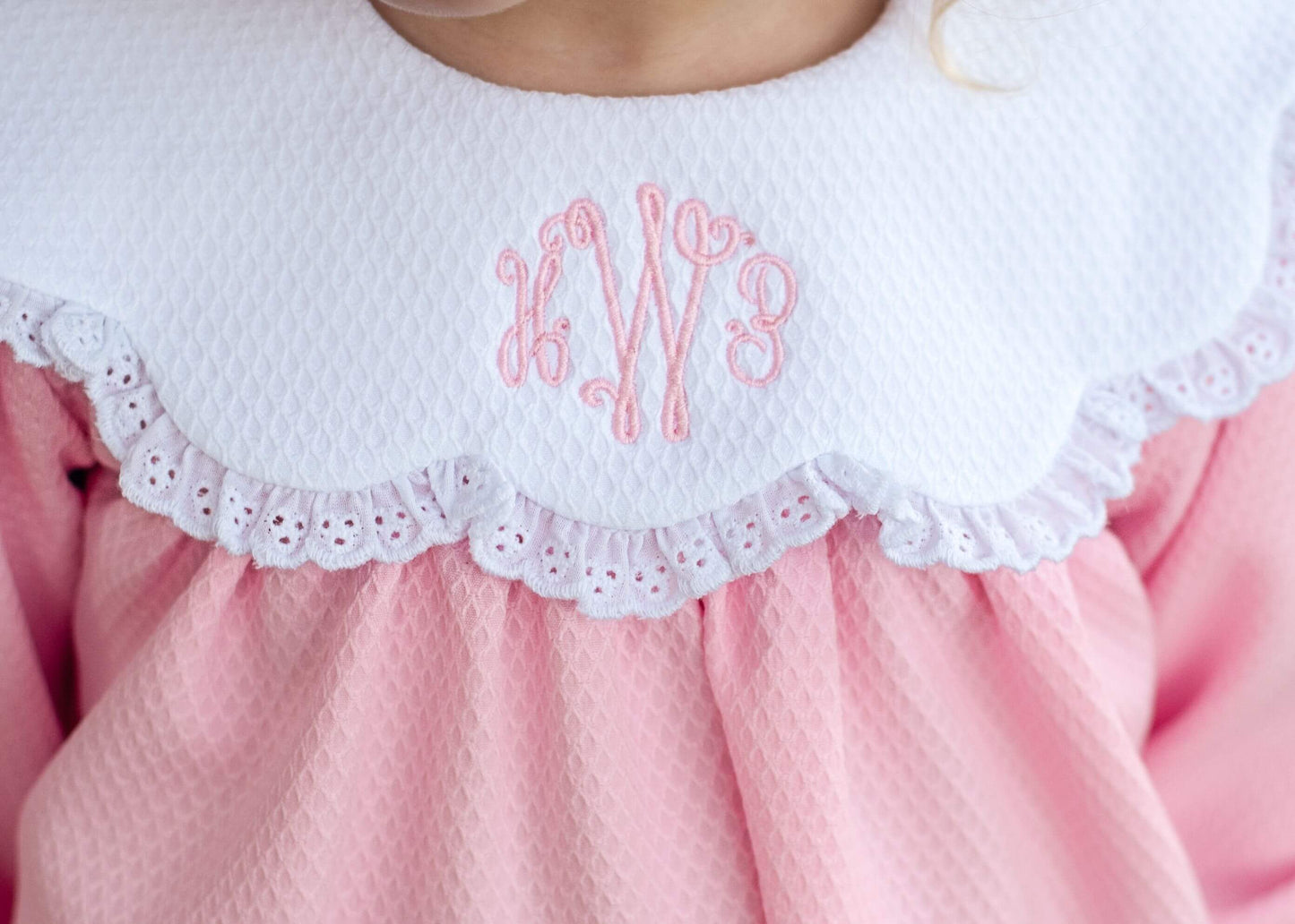 Close-up of a child wearing a pink dress with a white ruffled eyelet collar featuring pink embroidered monogram letters.