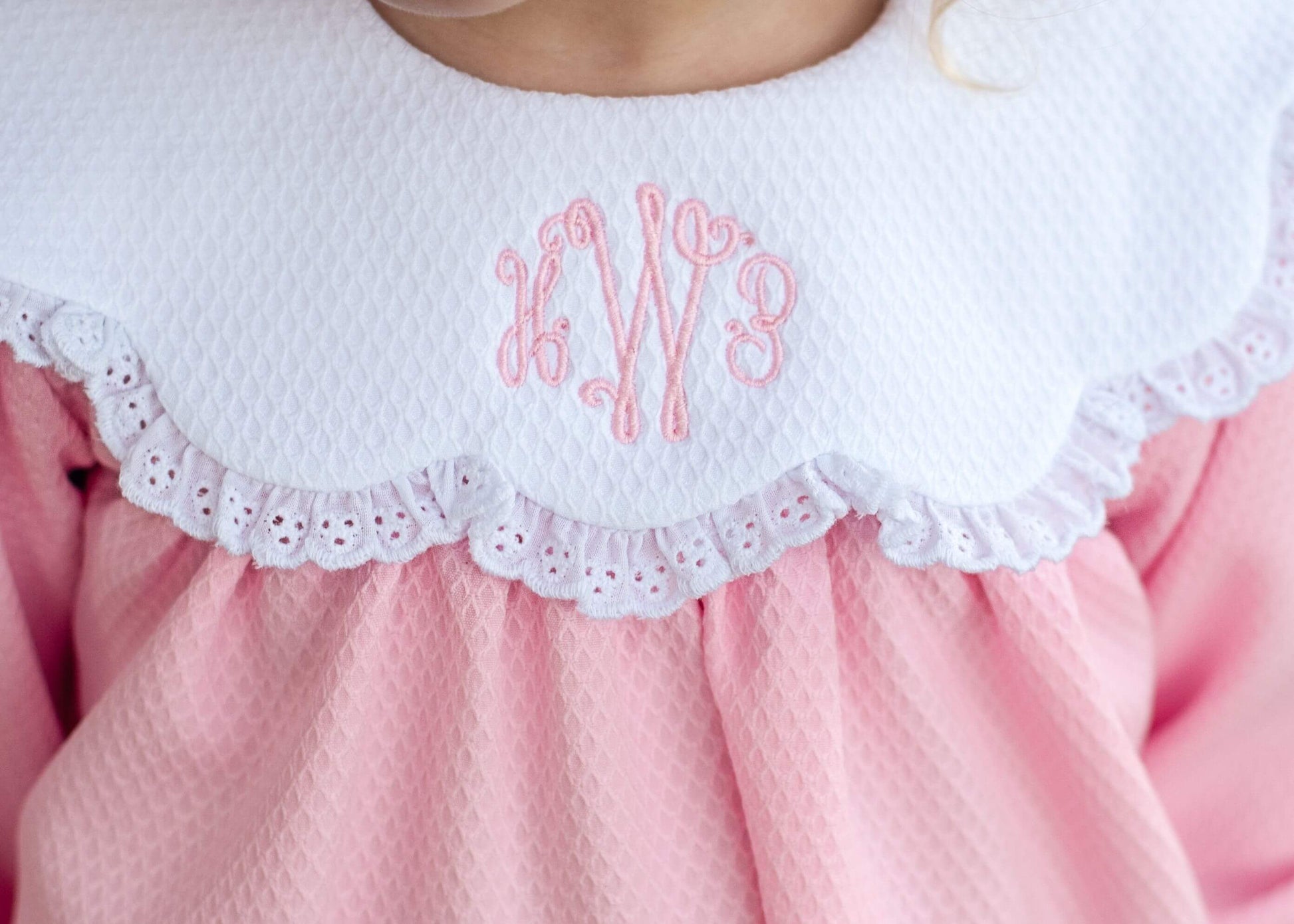Close-up of a child wearing a pink dress with a white ruffled eyelet collar featuring pink embroidered monogram letters.