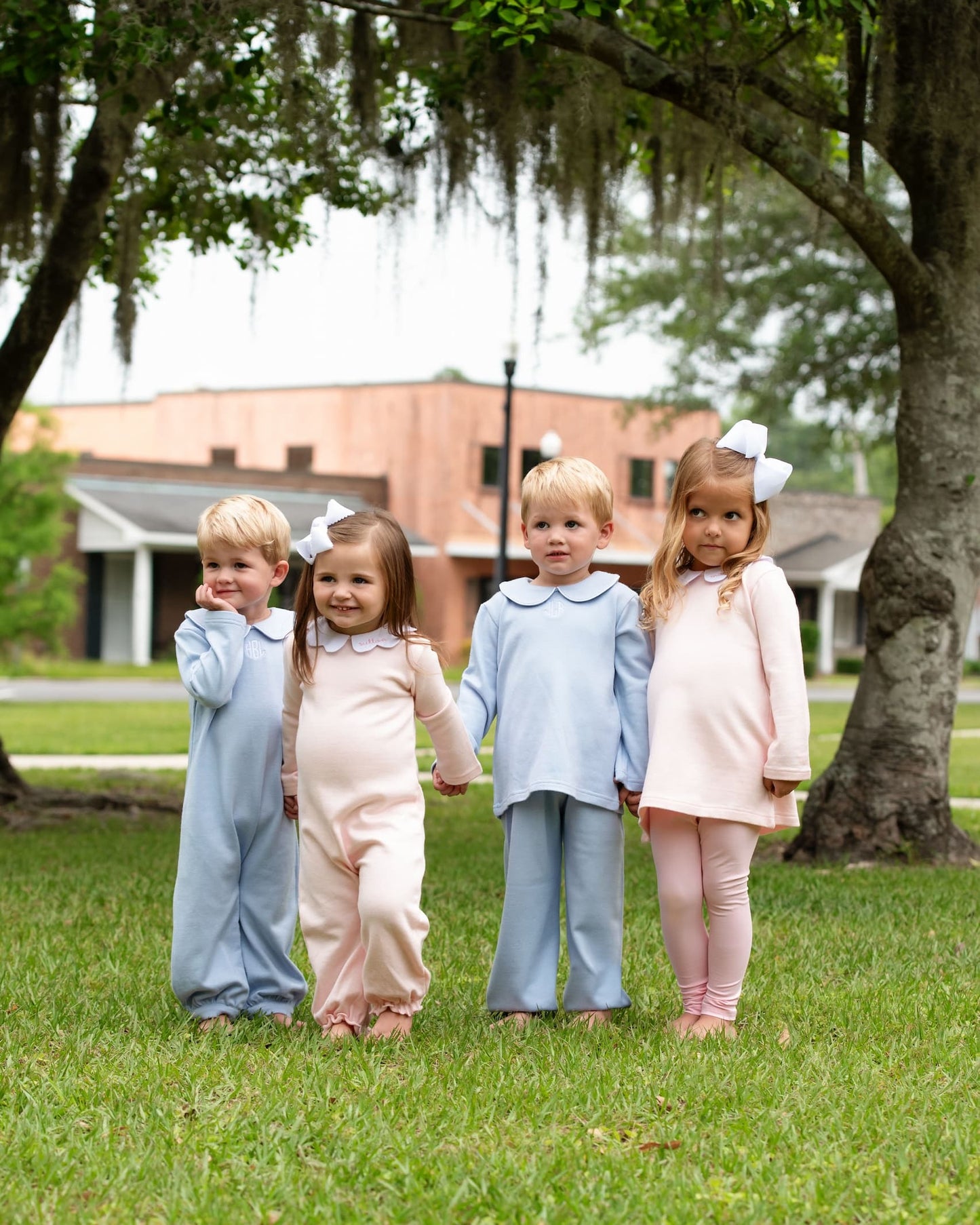 Four children in matching outfits standing in a grassy area with trees and a building in the background.
