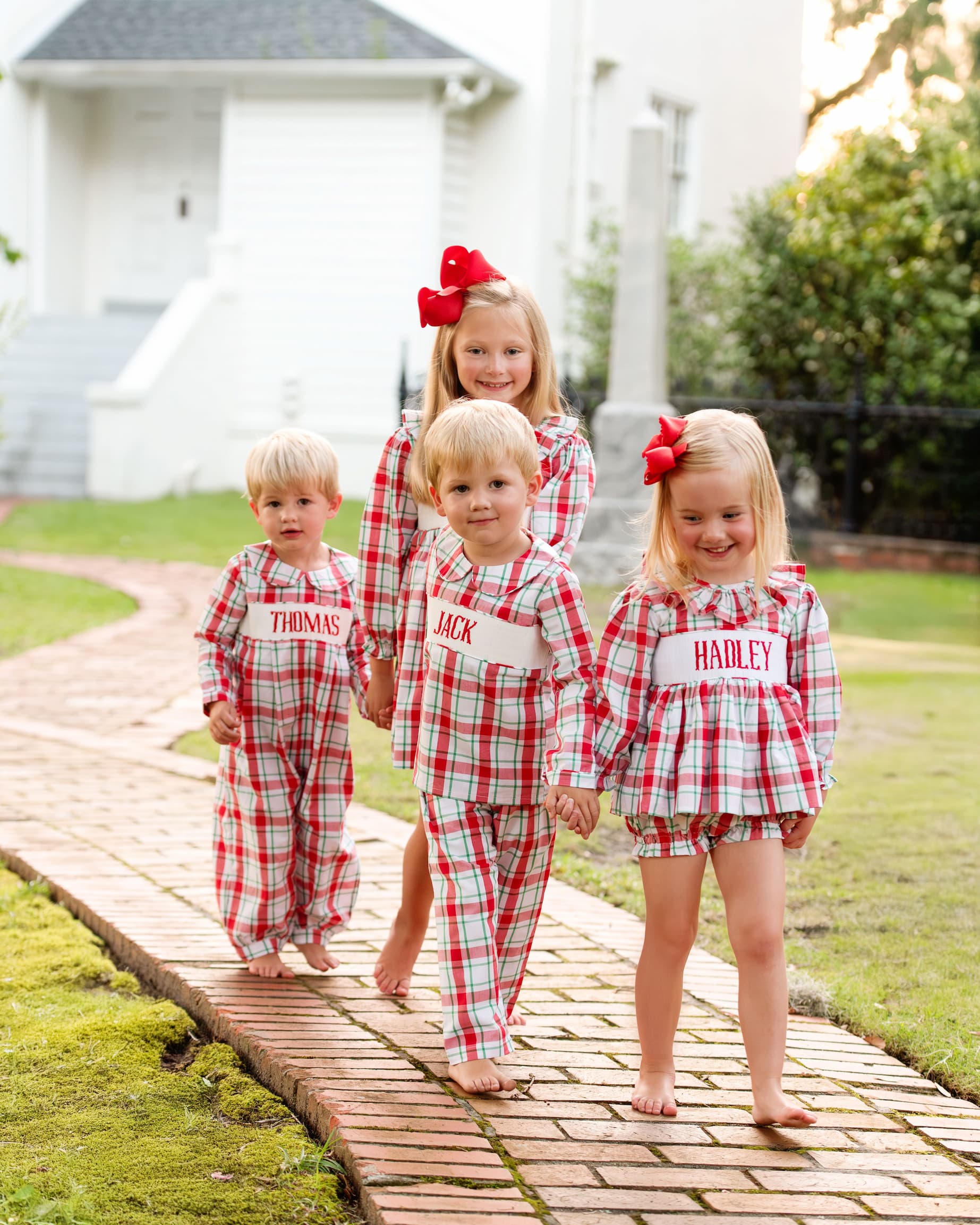 Four children in matching red and white checkered outfits standing on a wooden path outdoors.