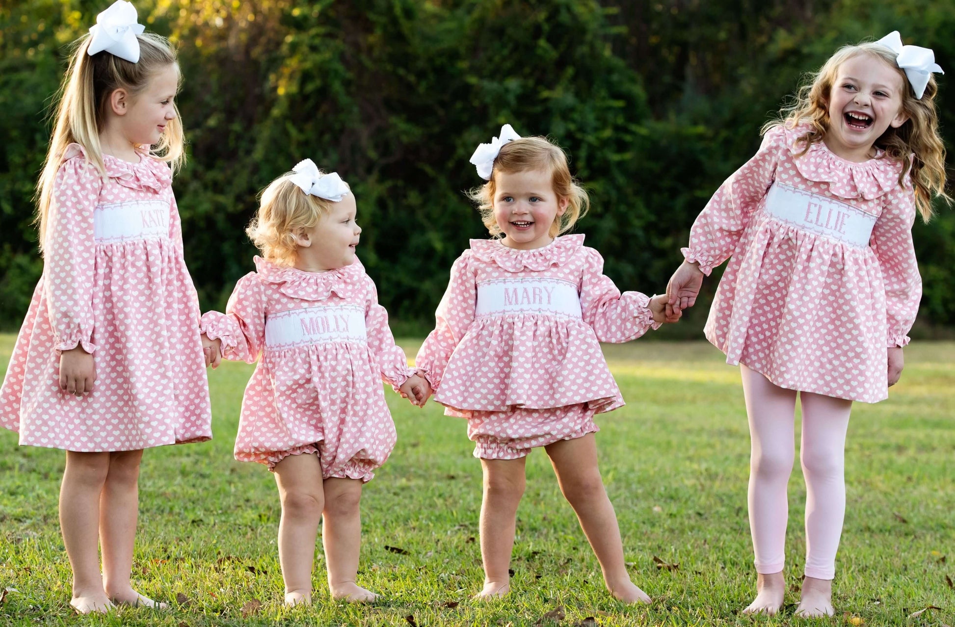 Four young girls in matching pink dresses holding hands outdoors.