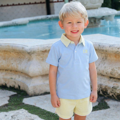 Young boy in a light blue shirt and yellow shorts standing in front of a fountain.