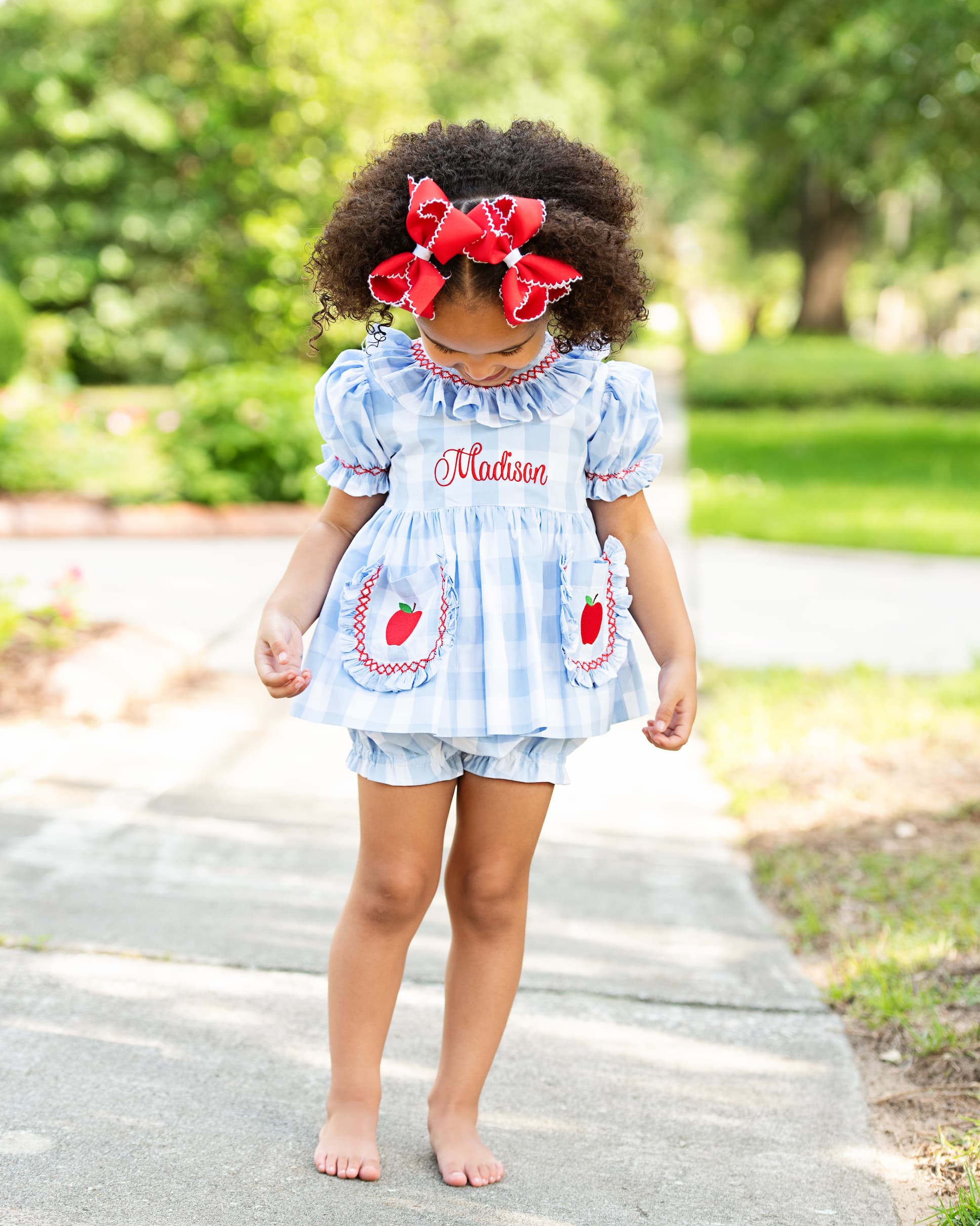Girl barefoot walking on a sidewalk wearing a blue outfit with apples on the pockets.