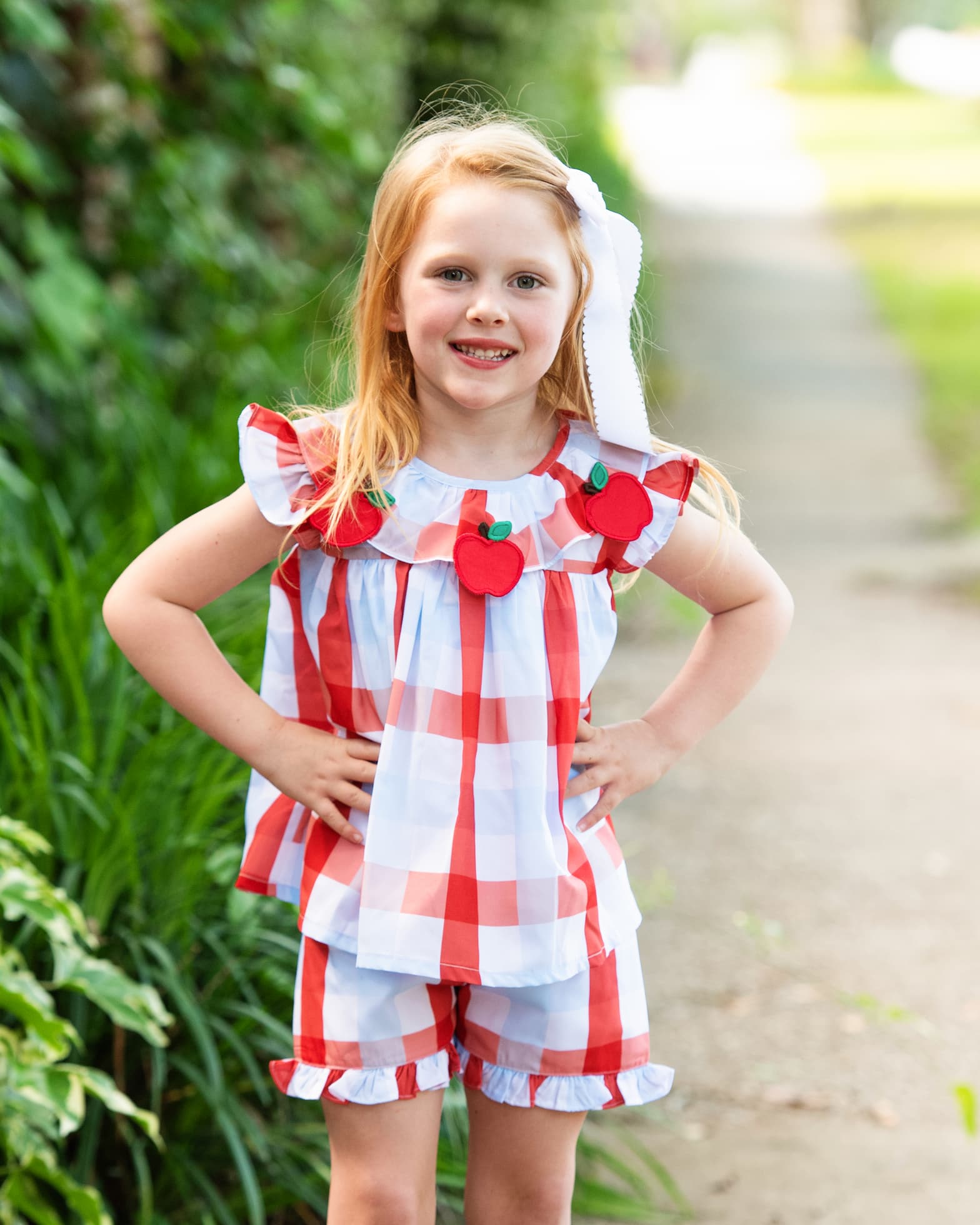 Girl in a plaid outfit with apples on it standing near bushes and a sidewalk.