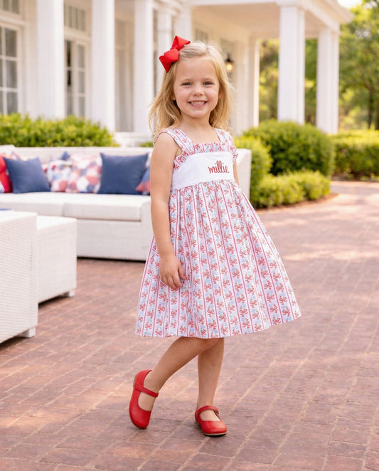 Girl in red and blue dress with name millie embroidered wearing a red bow and red shoes. 