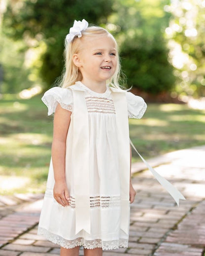 Little girl wearing an ivory heirloom christening dress with lace trim, flutter sleeves, and satin bows, standing outdoors for a special occasion portrait.