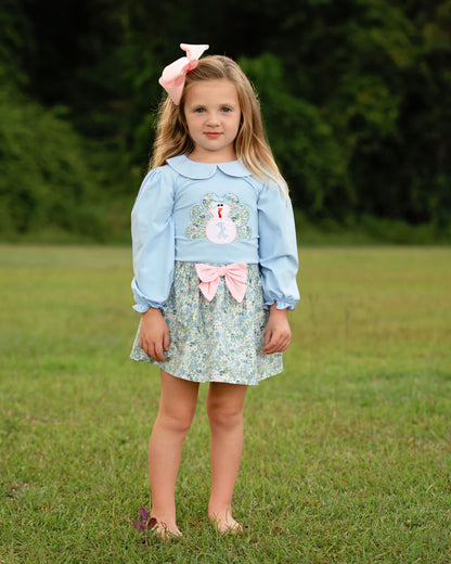 Girl standing barefoot on the grass wearing a floral skirt and a blue shirt with a turkey on it