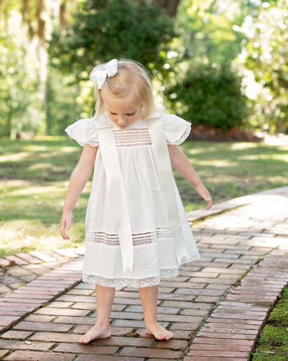 Little girl wearing an ivory heirloom christening dress with lace panels, flutter sleeves, and satin ribbon bows, standing barefoot on a brick path during a sunny outdoor photo session.
