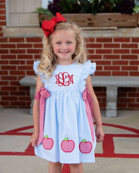 Girl wearing a blue dress with apples and a monogram at the chest.