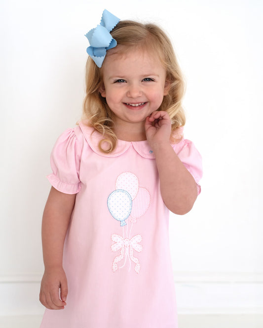 Girl wearing a dress with balloons and a bow on it smiling. 