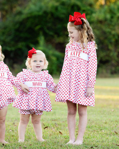 Three young girls in matching red and white heart dresses with smocked names outdoors. 