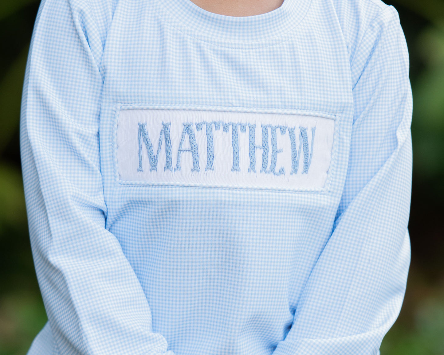 Light blue checkered shirt with 'MATTHEW' embroidered on the chest against a blurred green background