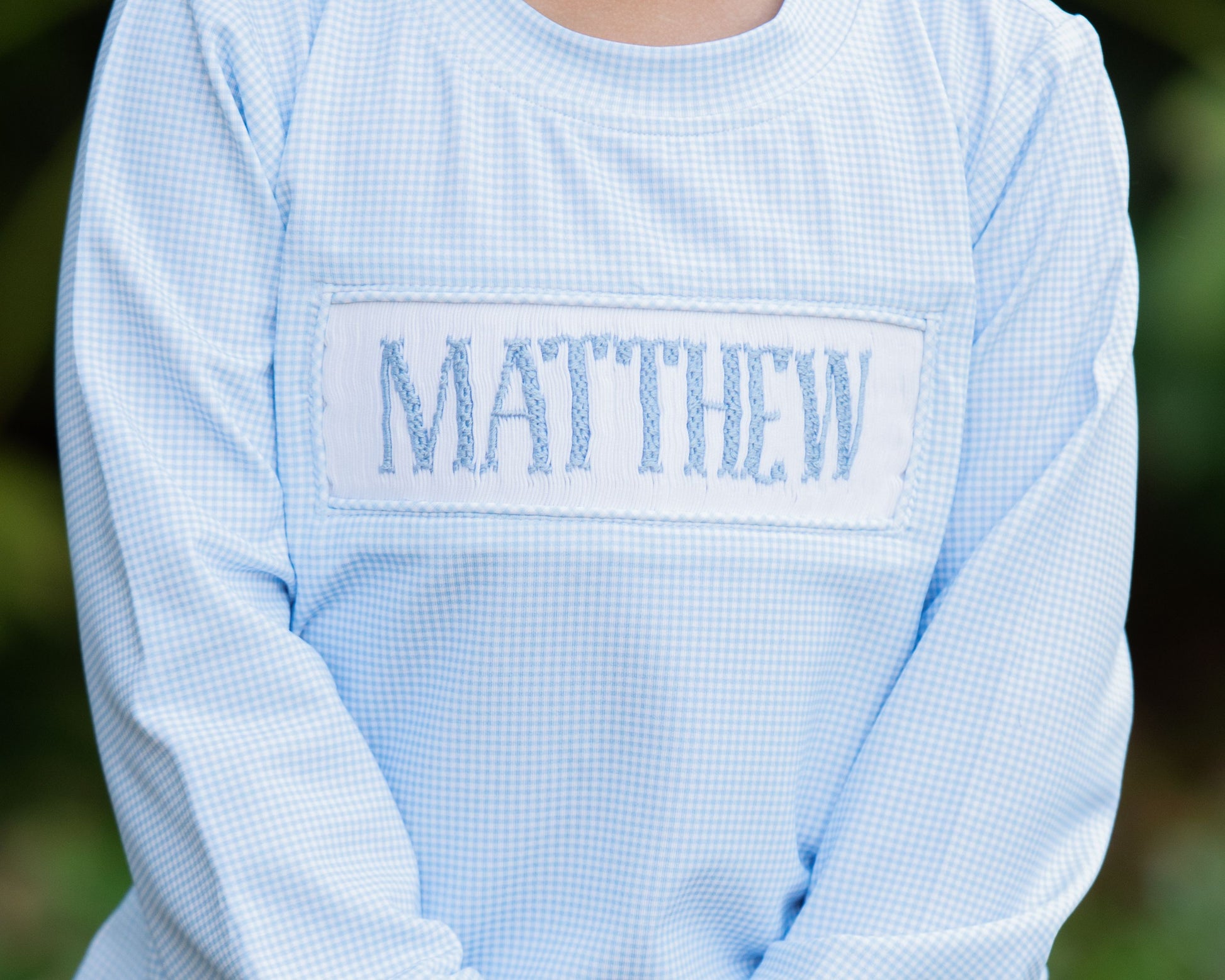 Light blue checkered shirt with 'MATTHEW' embroidered on the chest against a blurred green background