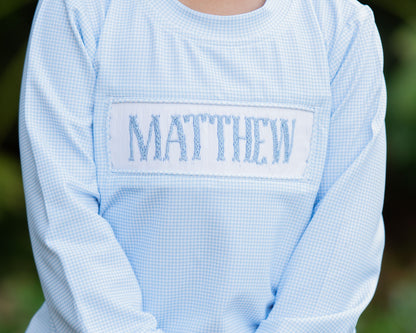 Light blue checkered shirt with 'MATTHEW' embroidered on the chest against a blurred green background