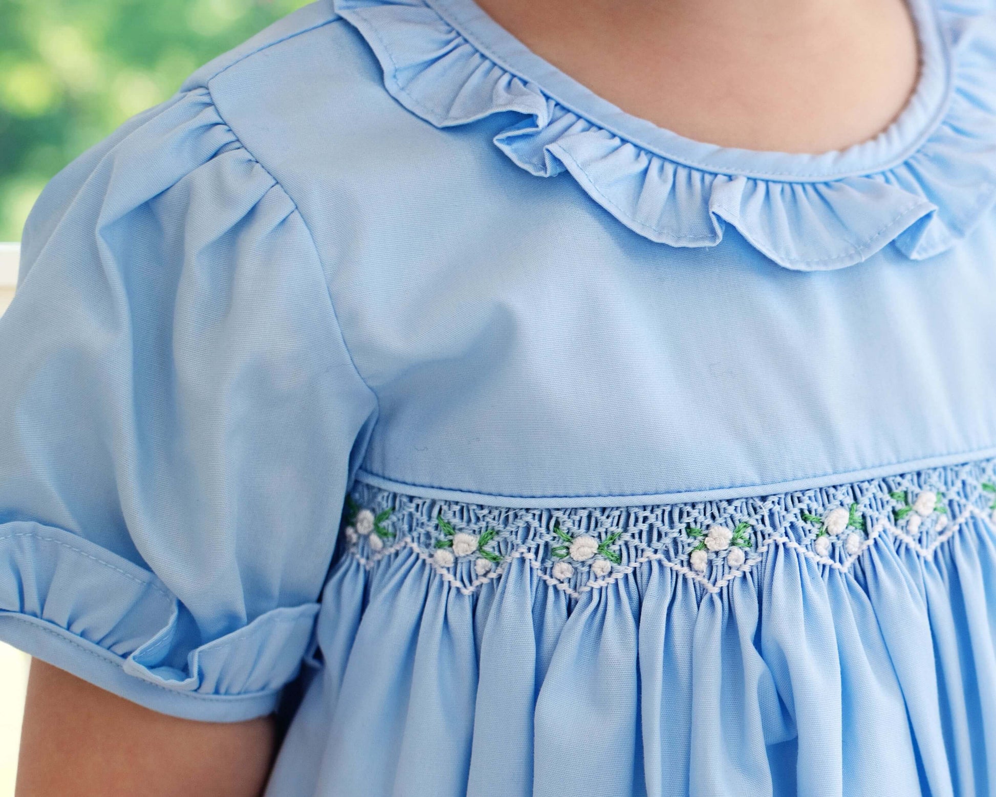 Light blue dress with ruffled collar and floral smocked embroidery on a blurred green background
