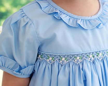 Light blue dress with ruffled collar and floral smocked embroidery on a blurred green background