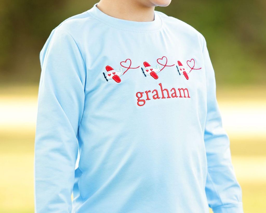 Light blue shirt with red footprints and hearts design and name 'graham' on a blurred natural background