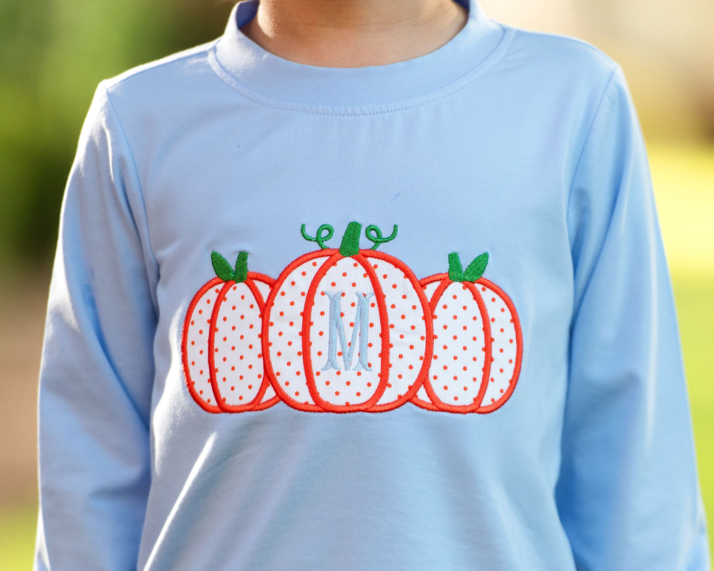 Light blue sweatshirt with pumpkin design and monogram 'M' on a blurred outdoor background