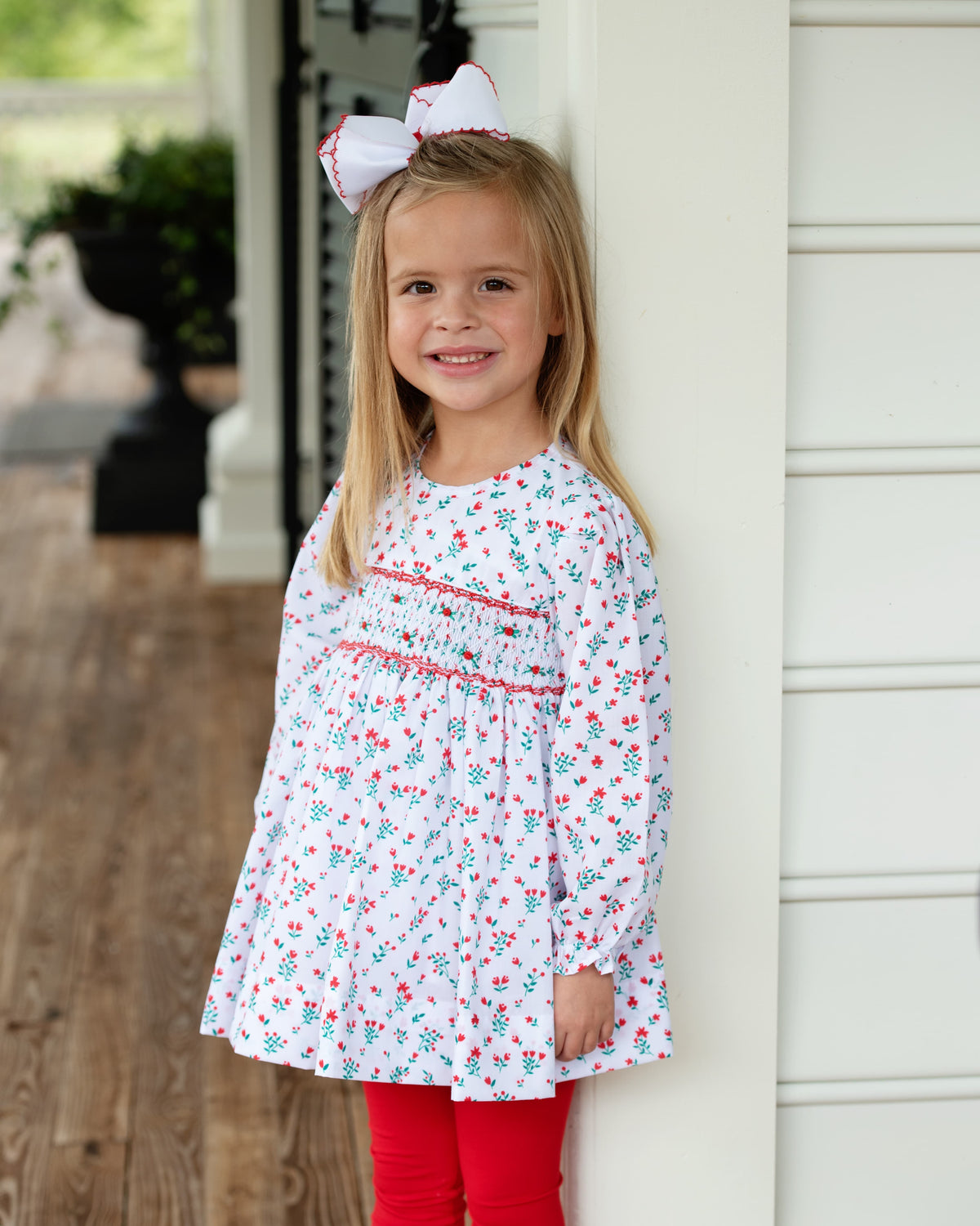 Little girl standing outside modeling a red and green floral smocked top with matching red leggings and a big bow in her hair