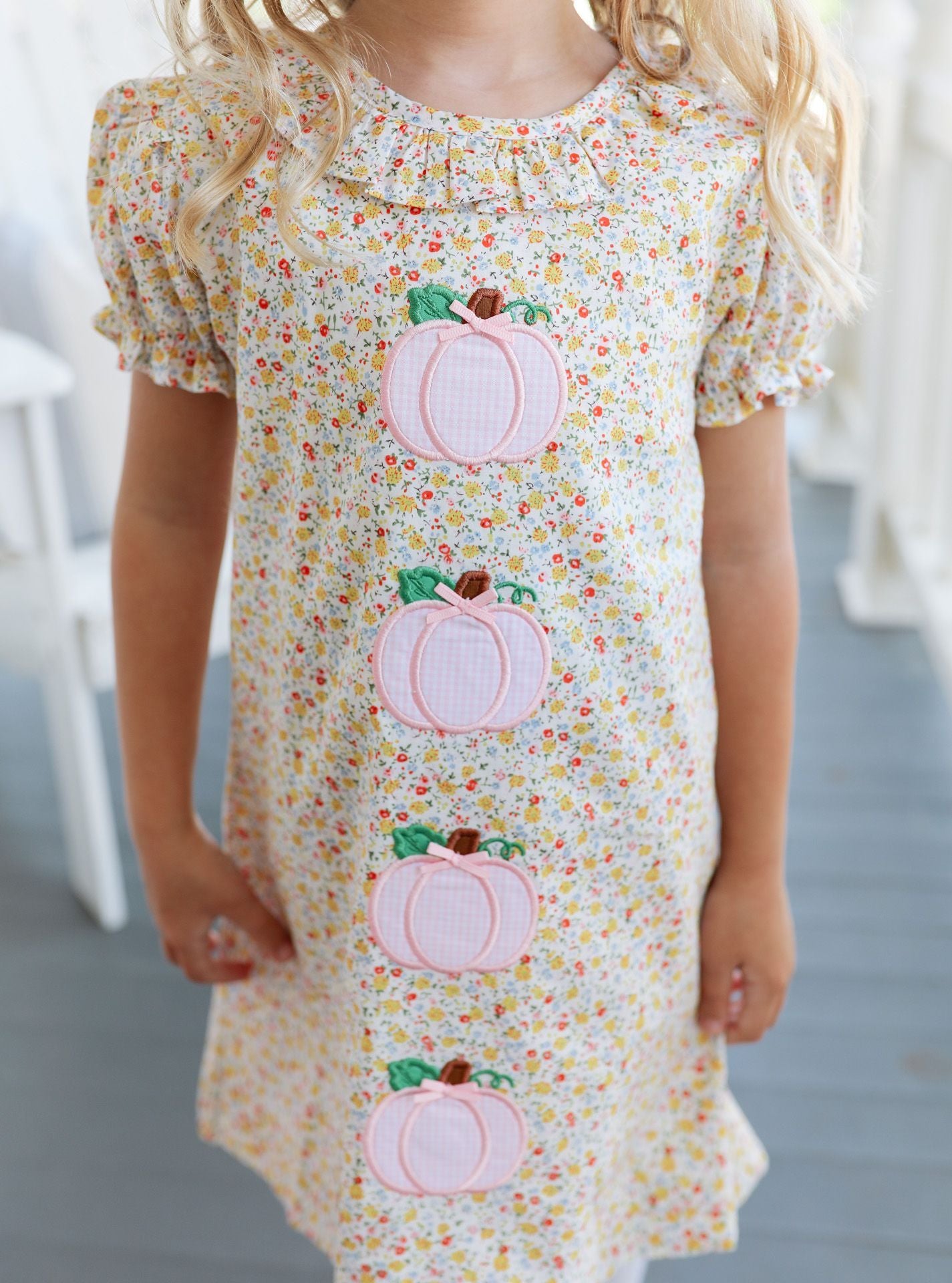 Child wearing a floral dress with pink pumpkin appliqués on a neutral background