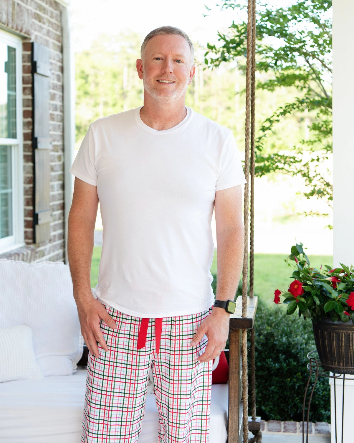 Man wearing a white t-shirt and red plaid pants standing on a porch.