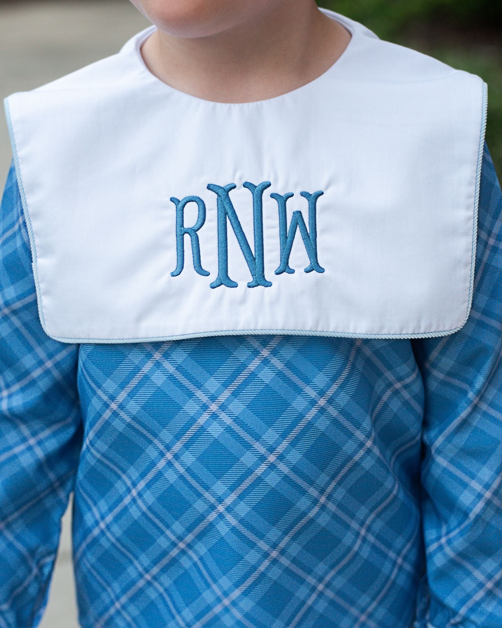 Child wearing a white bib with blue monogrammed letters over a blue plaid shirt.