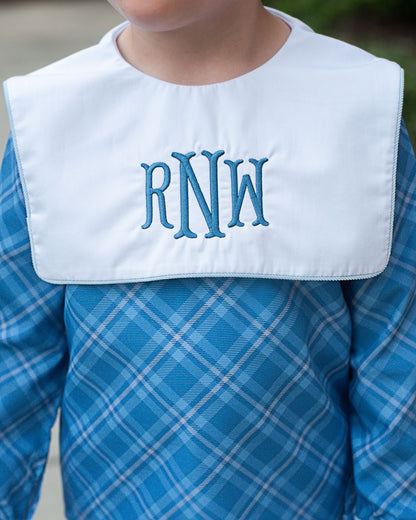 Child wearing a white bib with blue monogrammed letters over a blue plaid shirt.