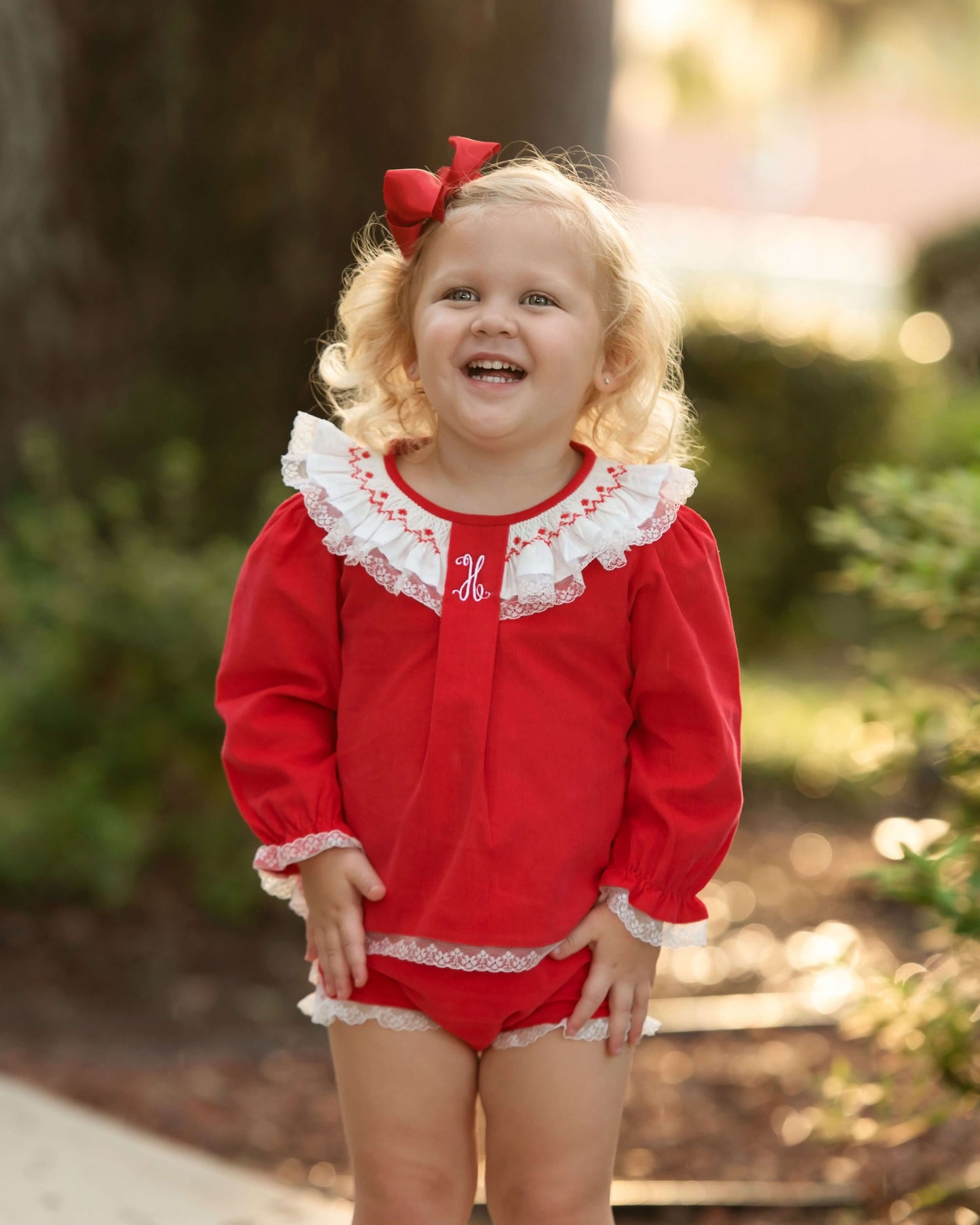 A young blonde girl wearing a red long-sleeve romper with white lace trim and a matching red bow in her hair, standing outdoors in a sunny, blurred park setting.
