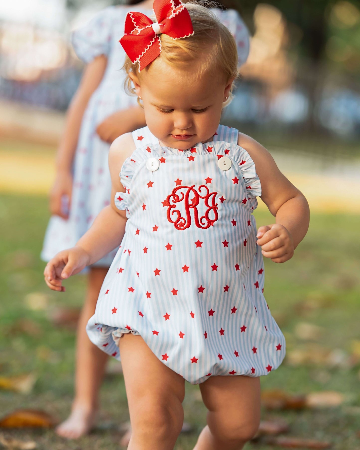 Small child wearing a star print outfit with a monogram on the chest and a red bow on her hair.